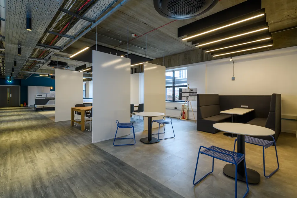 Modern office break area with partitioned tables and blue metal chairs under exposed ceiling with linear lighting.