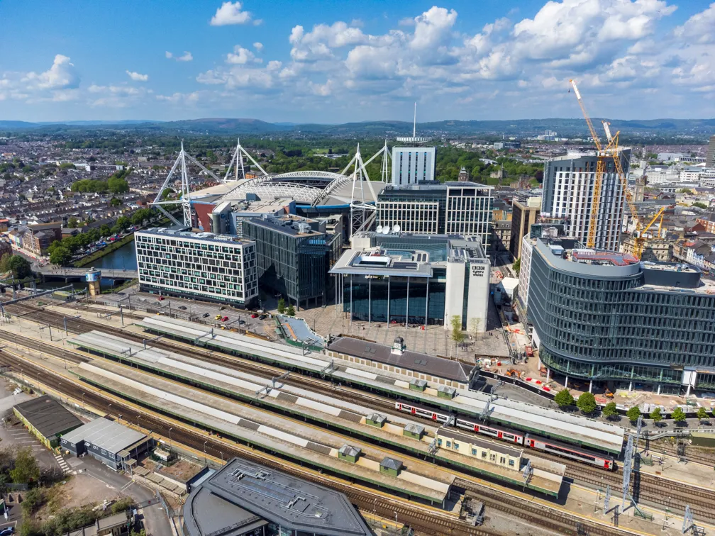 Aerial view of a large train station with multiple platforms and trains, surrounded by modern office buildings and a stadium with white support beams under a blue sky.