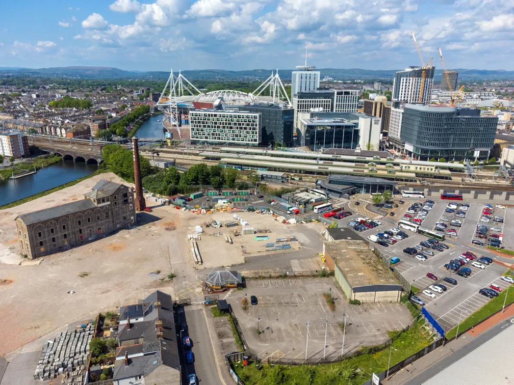 Aerial view of a cityscape featuring a large stadium with white support beams, a river with a stone bridge, railway tracks, parking lots, cranes, and surrounding buildings under a partly cloudy sky.