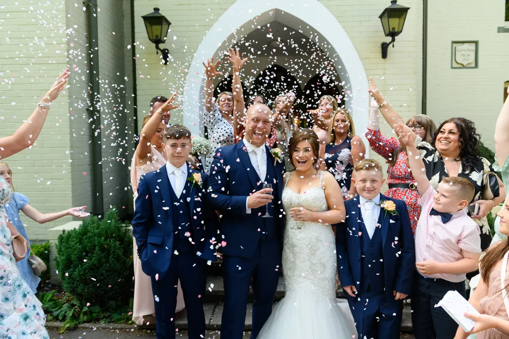 Bride and groom in wedding attire smiling and holding glasses amidst joyful guests throwing pink and white confetti.