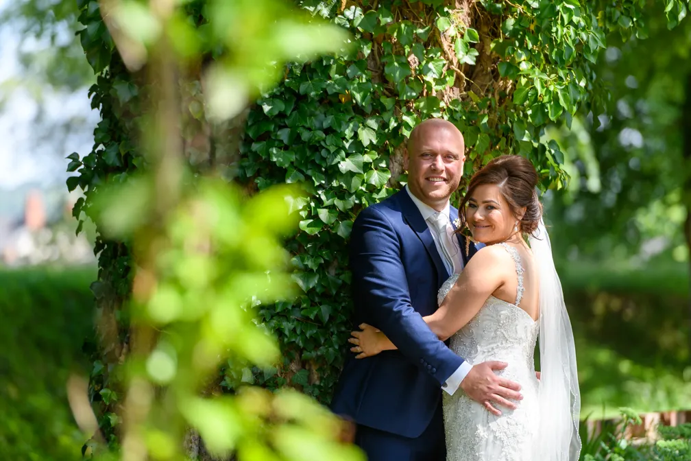 Bride and groom in wedding attire embracing and smiling outdoors next to a tree covered in green ivy.