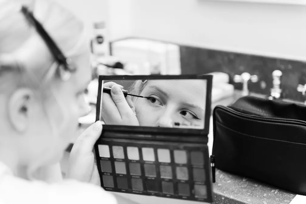 Woman applying eyeliner while looking into a palette mirror at a counter with a makeup bag nearby.