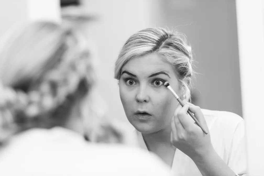 Woman applying eye makeup with a brush, looking into a mirror with a focused expression.