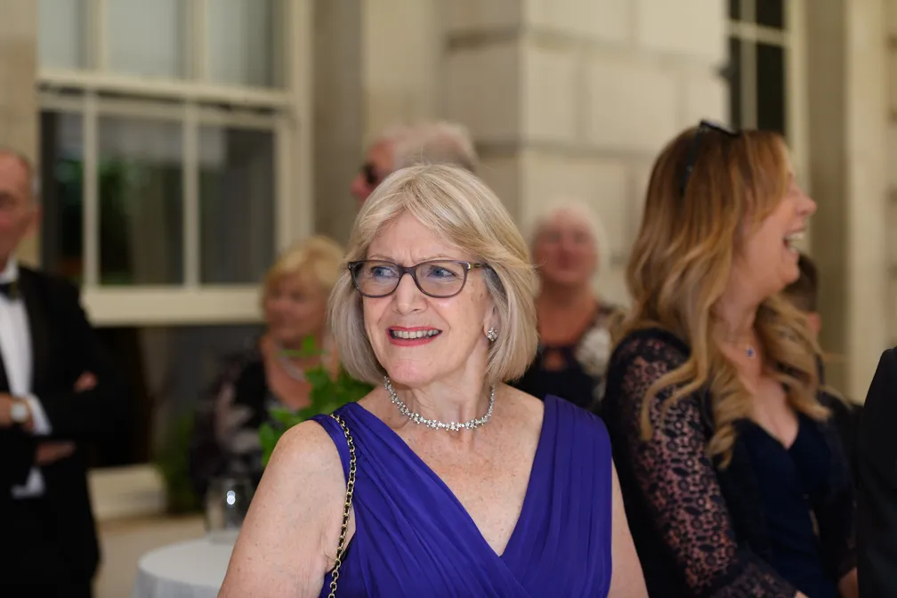 Smiling older woman with gray hair and glasses wearing a purple dress and a sparkly necklace at a social event.