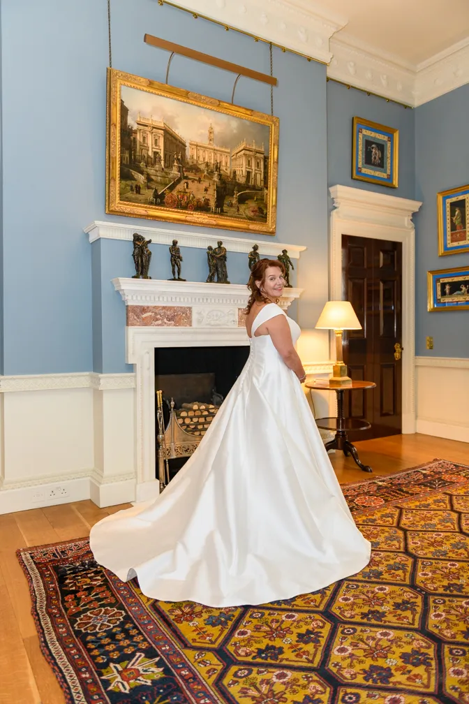 Bride in a white satin wedding gown smiling and standing in a blue-walled room with a fireplace, ornate paintings, and a patterned rug.