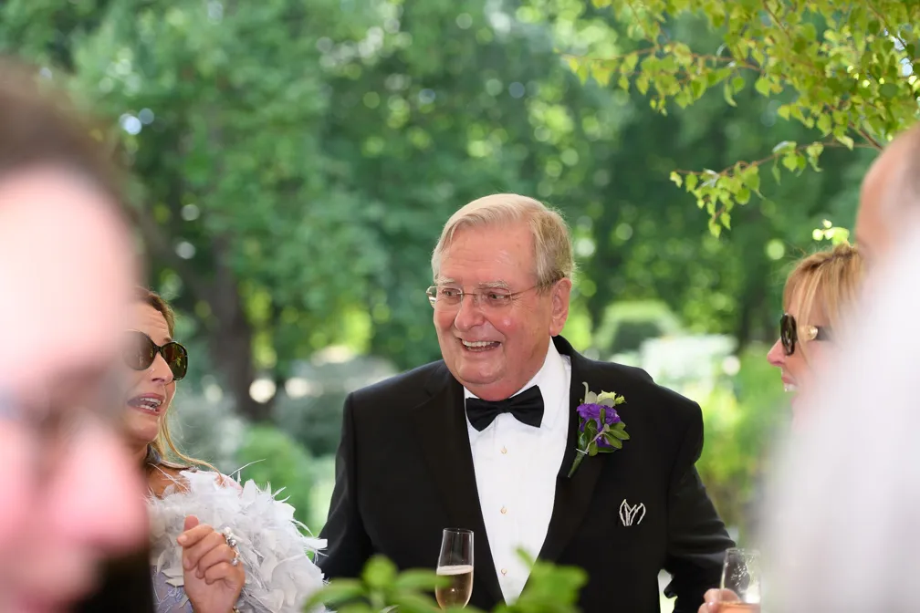 Older man in a black tuxedo with bow tie and purple boutonniere smiling and holding a glass of champagne at an outdoor gathering.