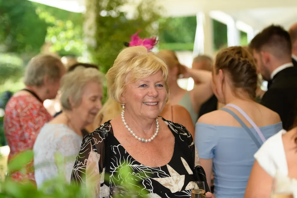Smiling elderly woman wearing a pearl necklace and black floral dress at a social gathering.