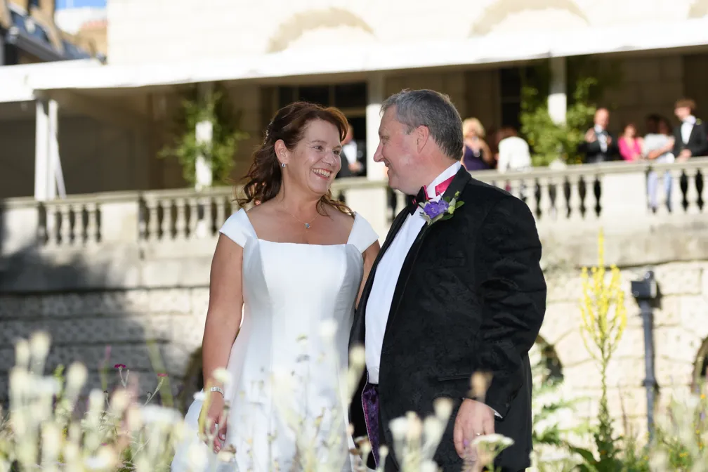Bride in white dress and groom in black tuxedo smiling at each other outdoors with stone building and guests in background.