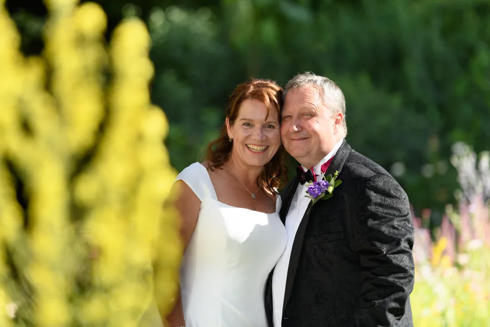 Smiling bride in white dress and groom in black tuxedo with purple boutonniere standing close together outdoors with greenery and yellow flowers in the background.