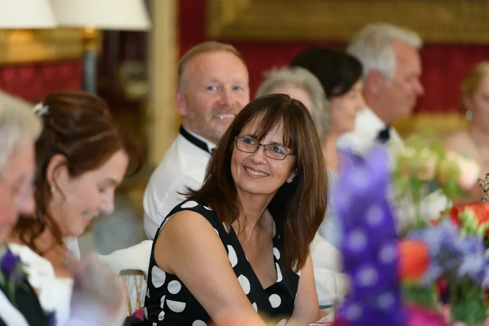 Smiling woman with glasses in a black dress with white polka dots sitting at a formal event table with other attendees.