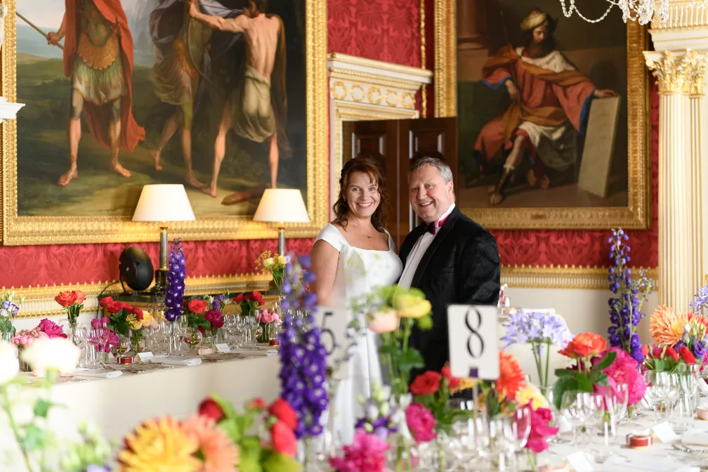 Smiling bride in a white dress and groom in a tuxedo standing together in an elegant room with red patterned walls, large classical paintings, and a dining table decorated with colorful flowers and place settings.