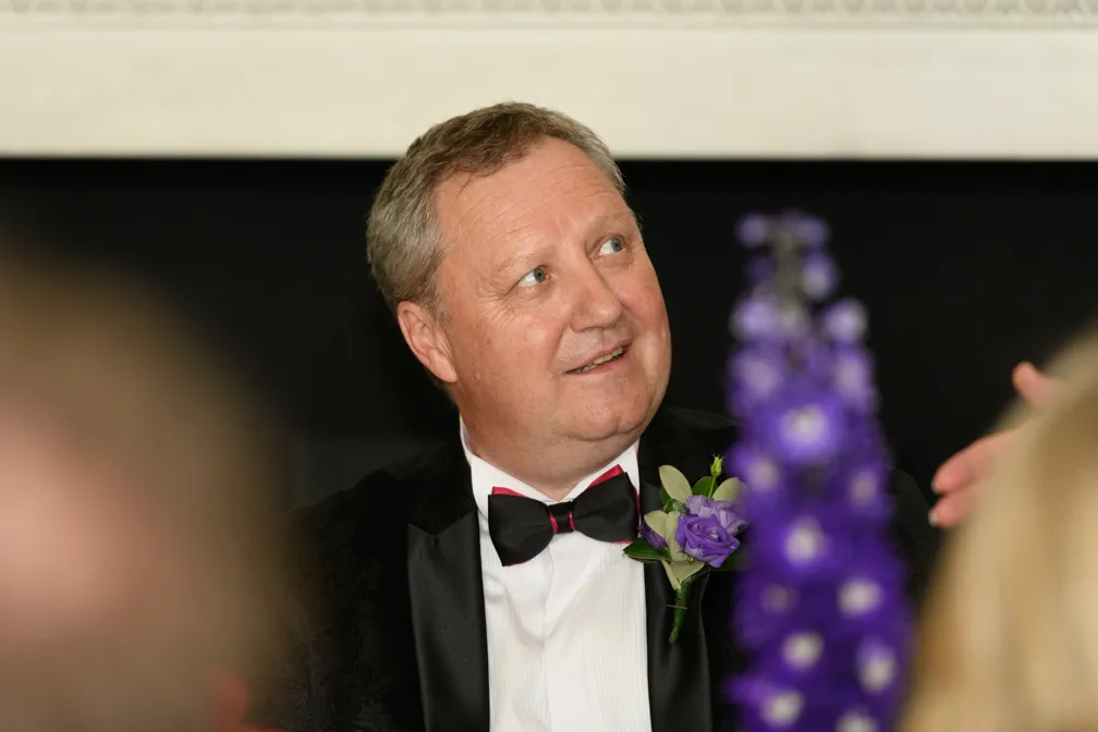 Middle-aged man in a black tuxedo with a bow tie and purple boutonniere looking to the side at an event.