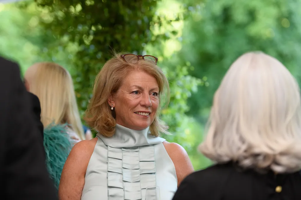 Smiling woman with blonde hair and glasses on her head, wearing a light blue ruffled sleeveless top, engaged in conversation with another person.