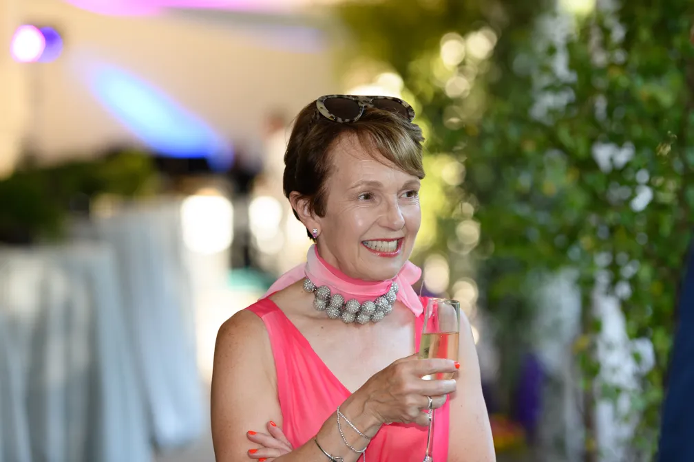 Smiling woman in a sleeveless pink dress holding a glass of champagne, wearing a beaded necklace and sunglasses on her head.
