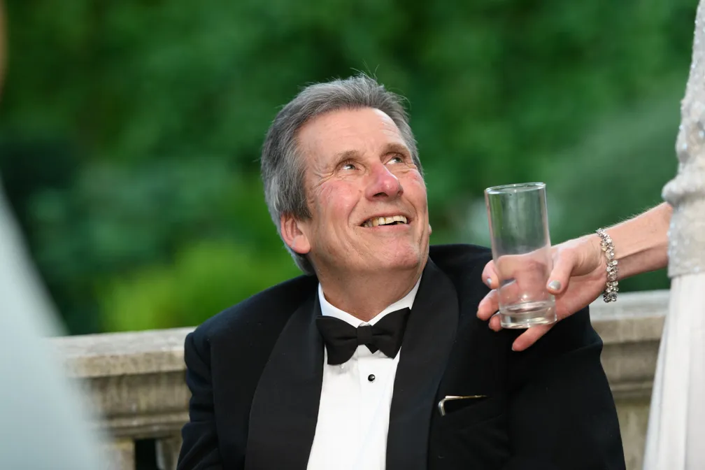 Smiling elderly man in a tuxedo looking up as a hand offers him an empty glass outdoors.
