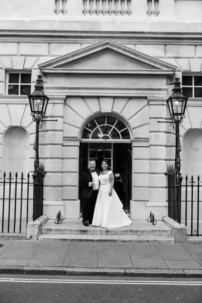 Bride in white gown and groom in tuxedo standing together in front of a grand stone building entrance.