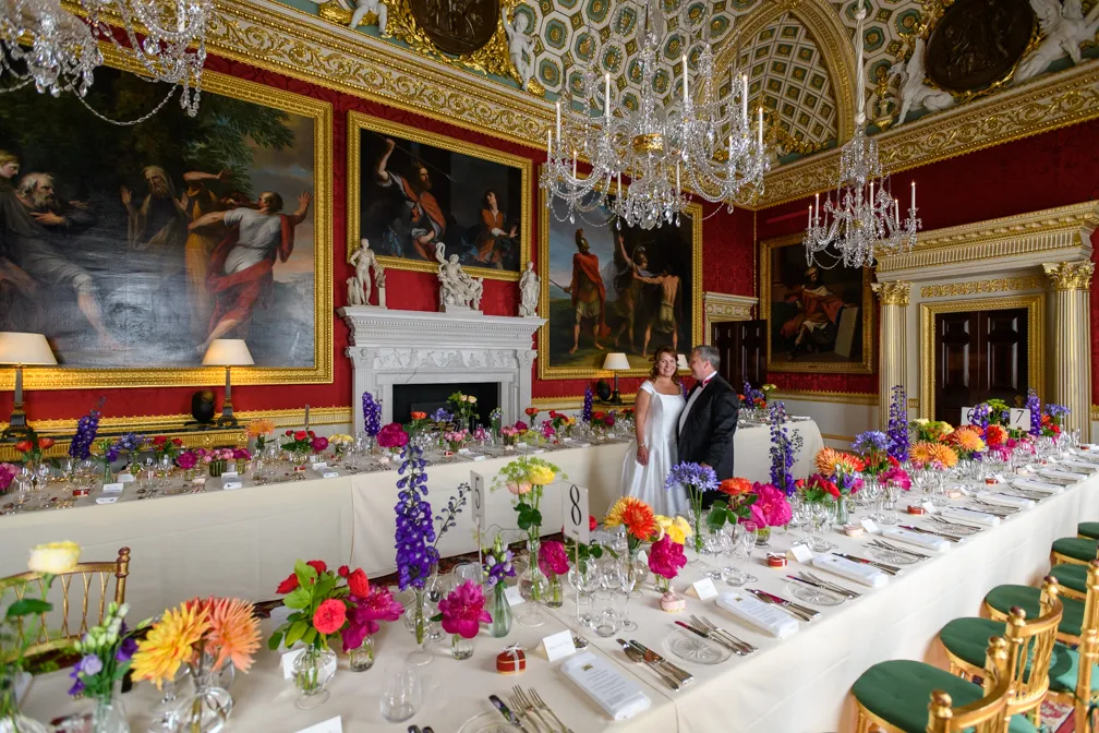 A bride and groom stand together smiling in an ornate, historic banquet hall with red wallpaper, large classical paintings, chandeliers, and long tables set with colorful flower arrangements and fine dining settings.