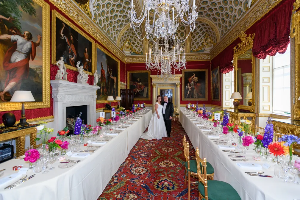 Bride and groom standing between two long banquet tables set with flowers and glassware in an ornate room with red walls, gold-framed paintings, and crystal chandeliers.