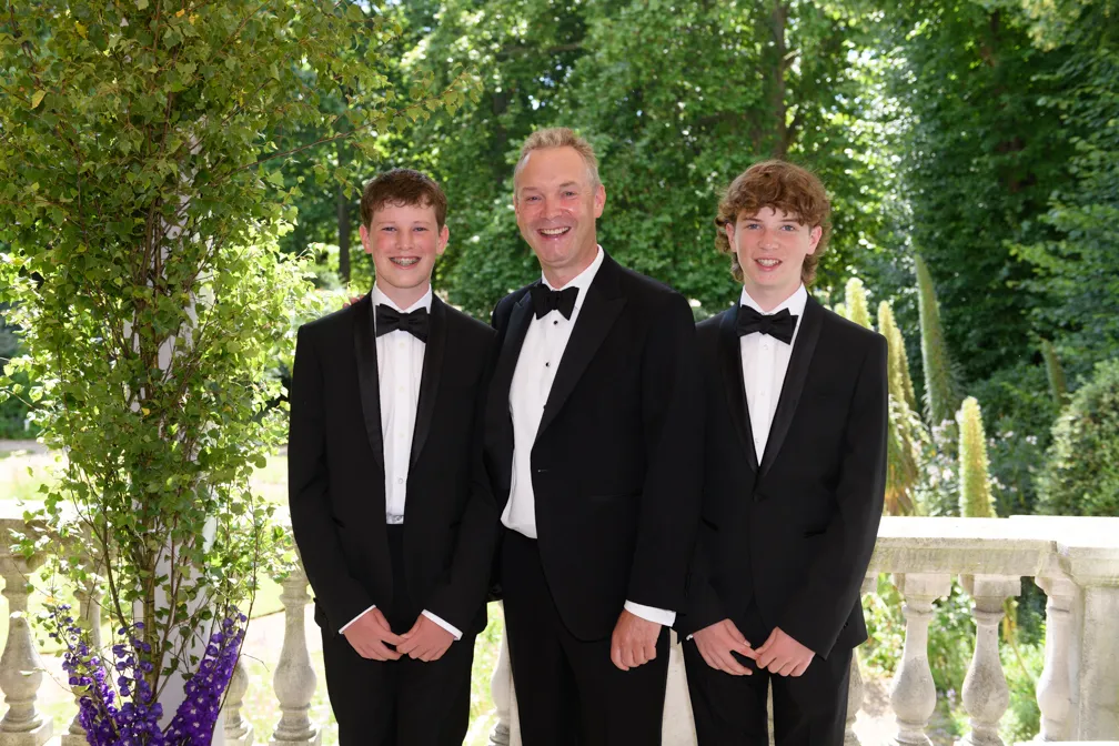 Three males in black tuxedos with bow ties smiling outdoors beside a stone railing and greenery.