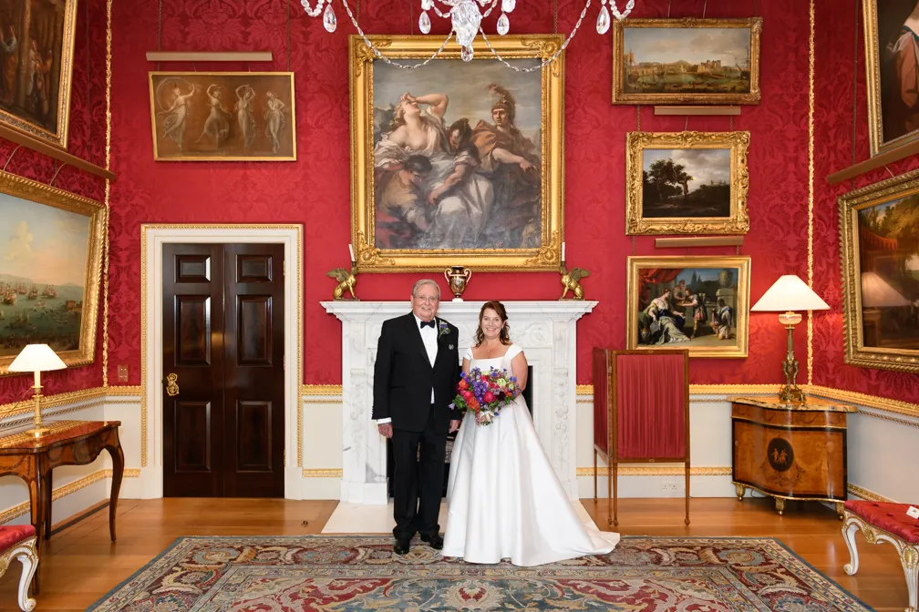 Bride in a white wedding gown holding a bouquet standing beside an older man in a black tuxedo in an ornate room with red wallpaper, classical paintings, and a white marble fireplace.