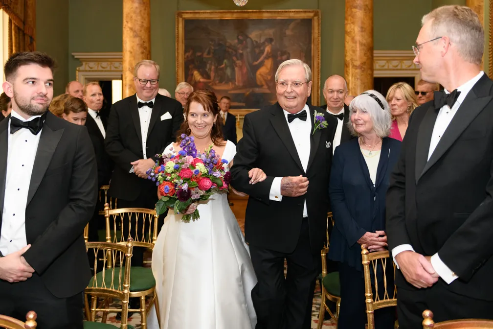 Bride in white gown holding a colorful bouquet walking arm-in-arm with an older man in a black tuxedo down an aisle lined with seated guests in formal attire.