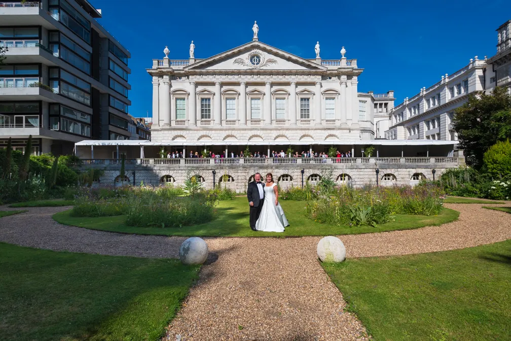 Bride and groom standing on a garden path with a historic white building and wedding guests on the balcony behind them under a clear blue sky.