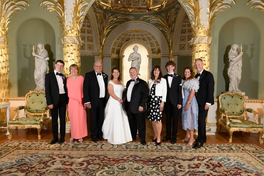 Group of people in formal attire standing in an ornate room with gold columns and classical statues, including a woman in a white dress and several men in tuxedos.