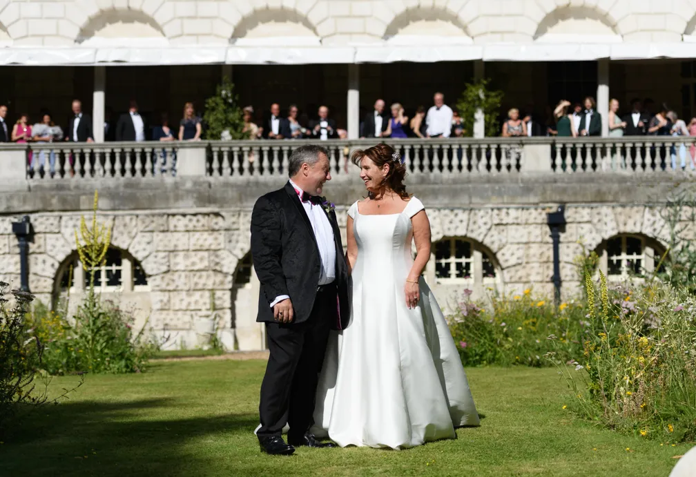 Bride and groom smiling and holding hands on a lawn in front of a stone building with spectators watching from a balcony above.