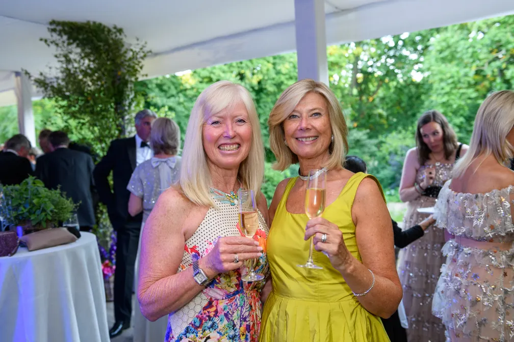 Two smiling women holding champagne glasses at an outdoor social event with other guests in the background.