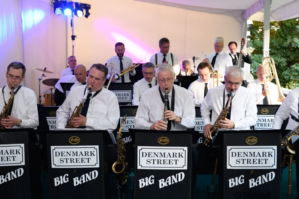 Denmark Street Big Band musicians performing with saxophones, clarinet, trombones, drums, and double bass under a tent.
