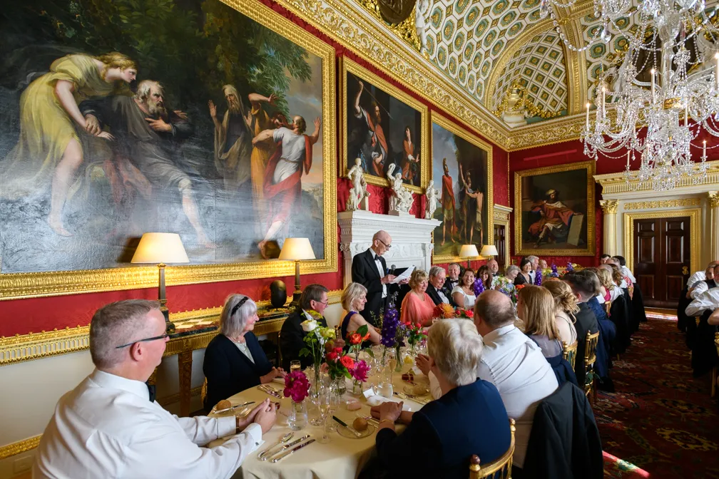 Group of formally dressed people seated at a long dining table in an ornate room with red walls, large classical paintings, and a crystal chandelier, listening to a man in a tuxedo standing and speaking.