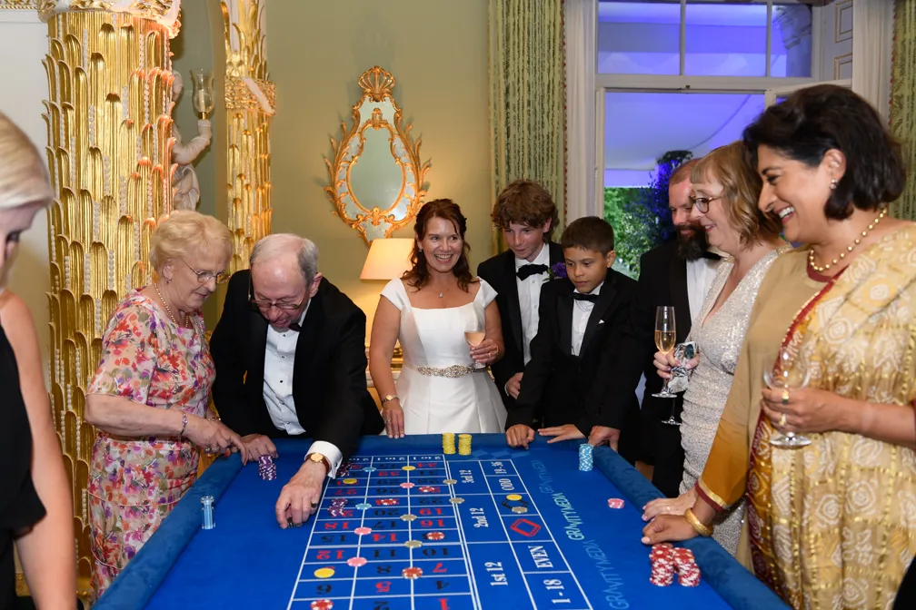 Group of people dressed formally playing casino roulette indoors around a blue felt table with chips and drinks.