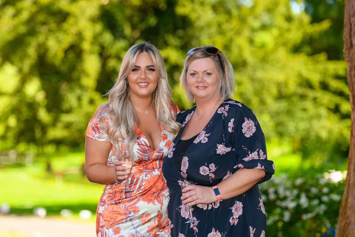 Two smiling women standing outside, one wearing a floral orange dress and the other a black floral dress, both holding glasses of champagne.