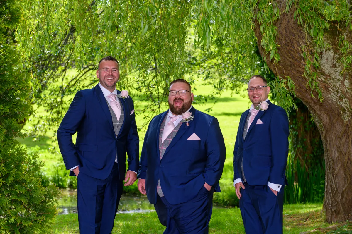 Three men in matching navy blue suits with pink ties and boutonnieres standing and smiling under a leafy tree in a green park.