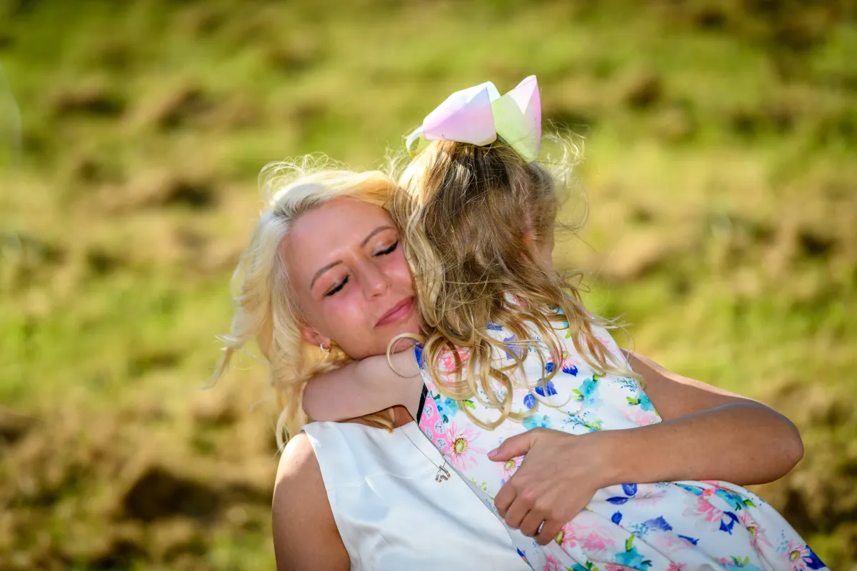 Blonde woman with eyes closed hugging a young girl wearing a floral dress and a large pastel bow in her hair outdoors.