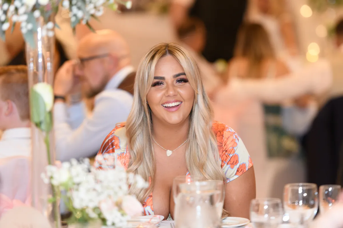 Smiling woman with long blonde hair wearing an orange floral dress seated at a dining table with glasses and flowers.