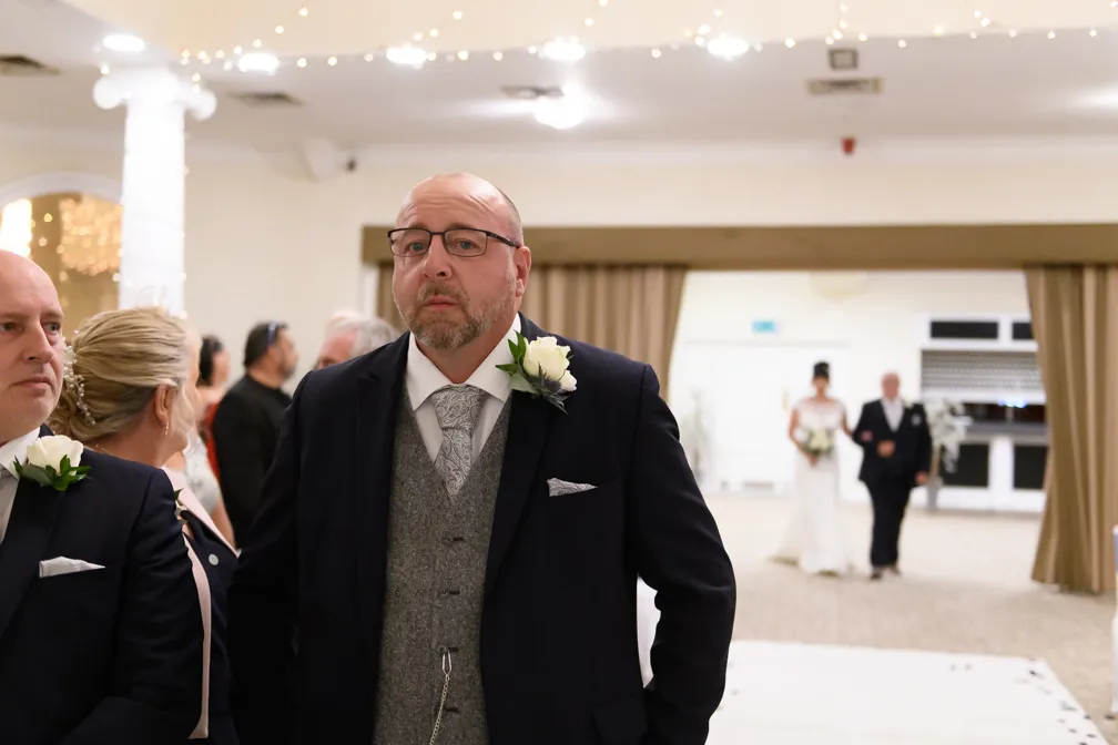 Man in glasses and formal suit with boutonniere looking ahead, with a bride and escort blurred in the background.