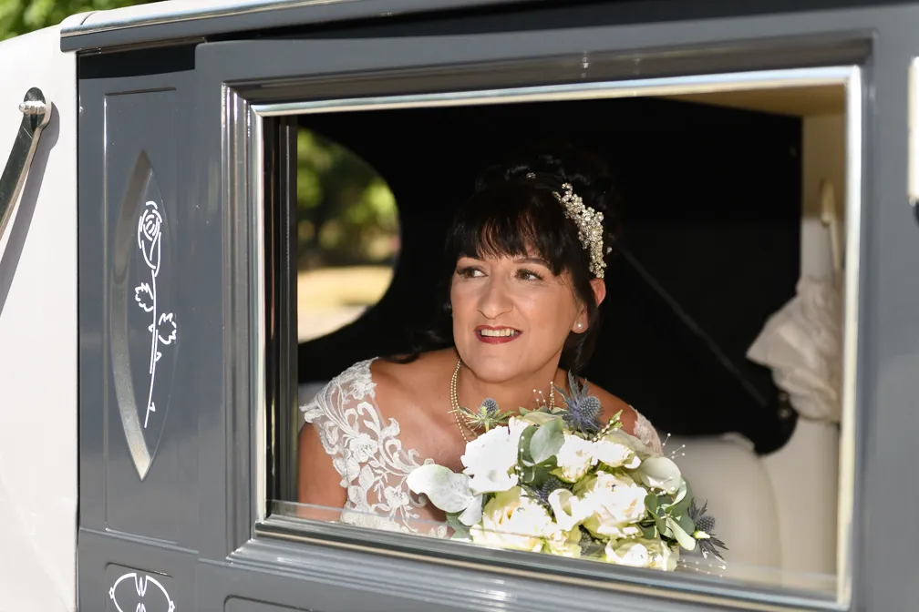 Smiling bride with dark hair, floral hairpiece, and lace wedding dress holding a bouquet while looking out of a car window.