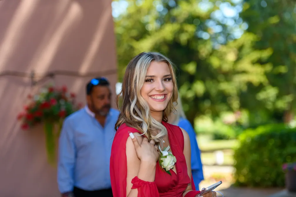 Smiling young woman in a red dress holding a phone outdoors with blurred people and greenery in the background.