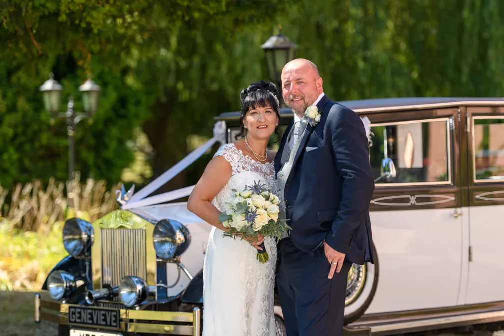 Bride in white lace wedding dress holding bouquet standing beside groom in dark suit in front of a classic white wedding car.