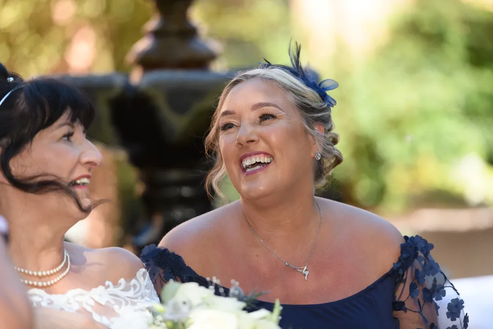 Two women smiling and laughing outdoors, one with a blue dress and floral headpiece, the other wearing a pearl necklace and white dress with lace.