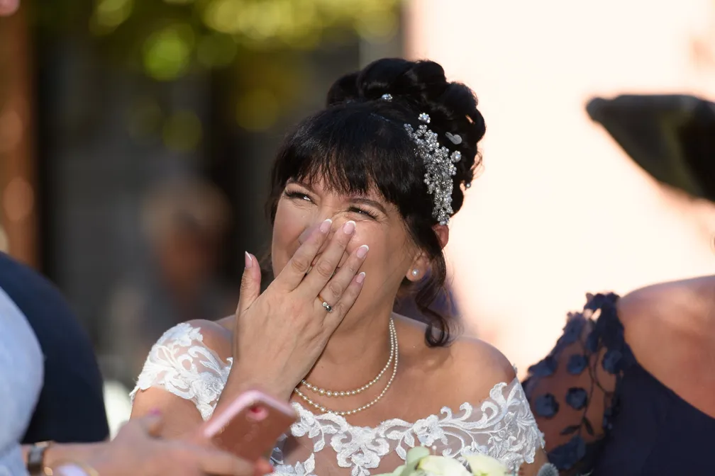 Bride in lace wedding dress with pearl necklace and jeweled hair accessory laughing and covering her mouth with one hand.