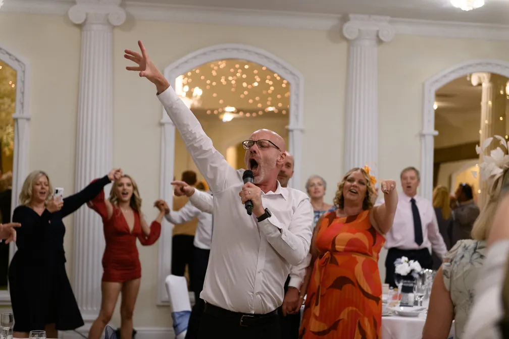 A man in glasses and white shirt singing enthusiastically into a microphone with one arm raised, surrounded by joyful people at a celebration.