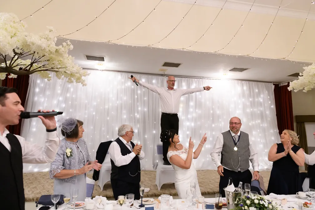 A lively wedding reception scene with a man standing on a chair holding a microphone and pointing, surrounded by smiling and clapping guests including the bride in a white dress.