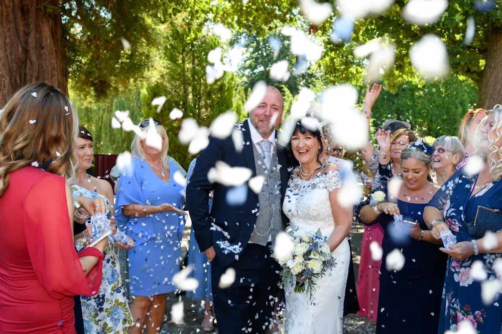 Bride and groom smiling amid guests throwing white flower petals outdoors during wedding celebration.