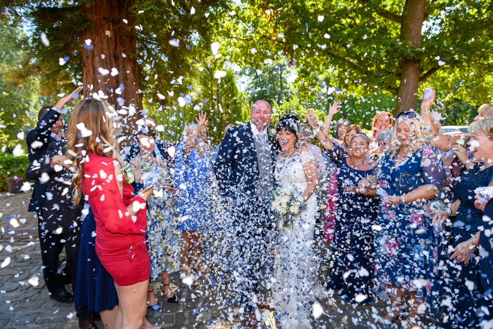 Bride and groom smiling while guests throw white flower petals during an outdoor wedding celebration under large green trees.