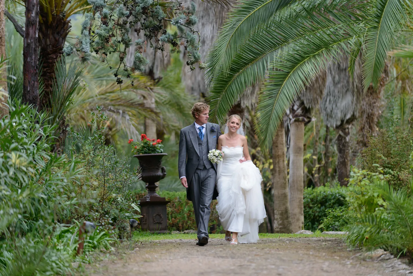 Bride in a white wedding dress holding a bouquet and groom in a gray suit walking arm in arm on a garden path lined with palm trees.