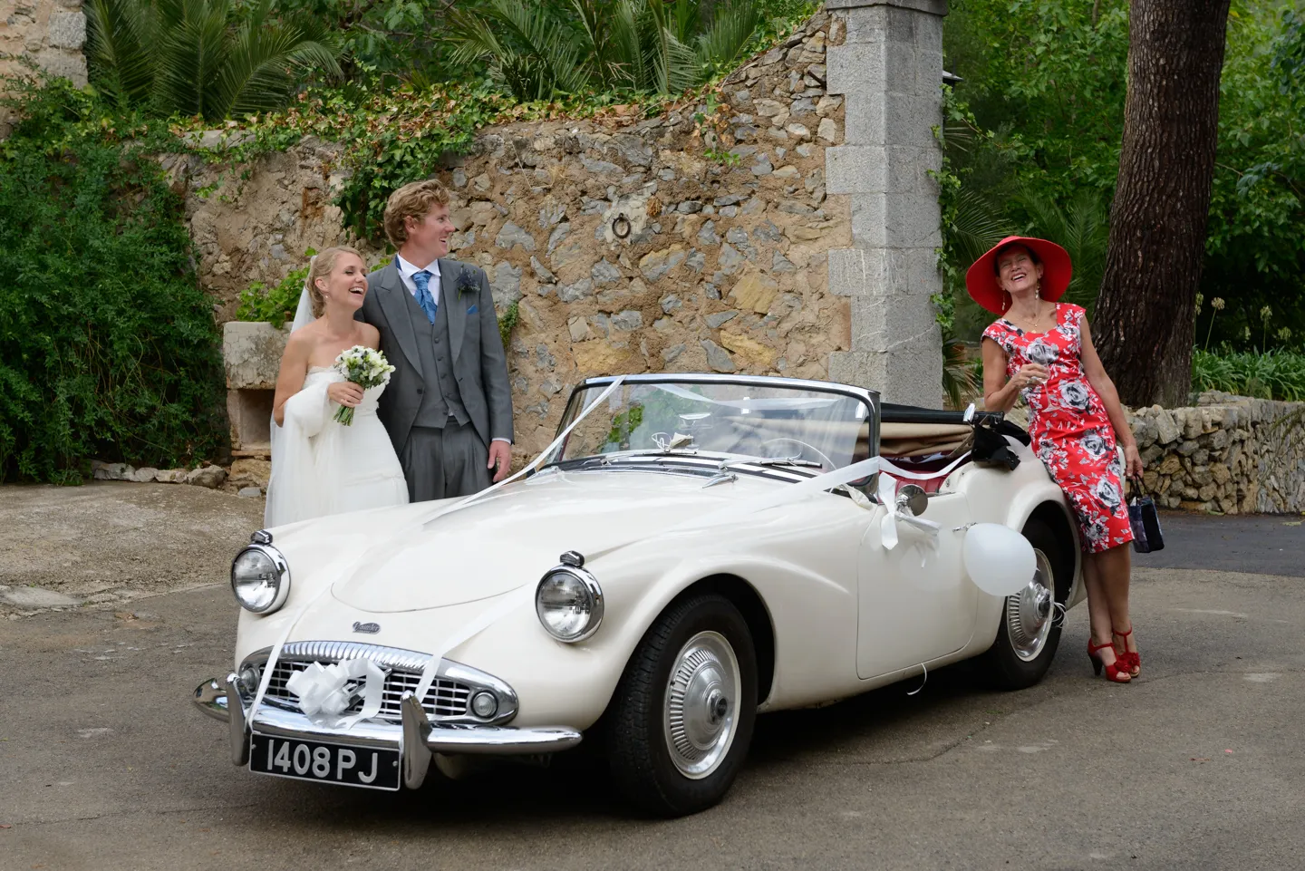 Bride and groom standing by a white vintage convertible car decorated with ribbons and balloons, with a woman in a red floral dress and wide-brimmed hat leaning on the car.