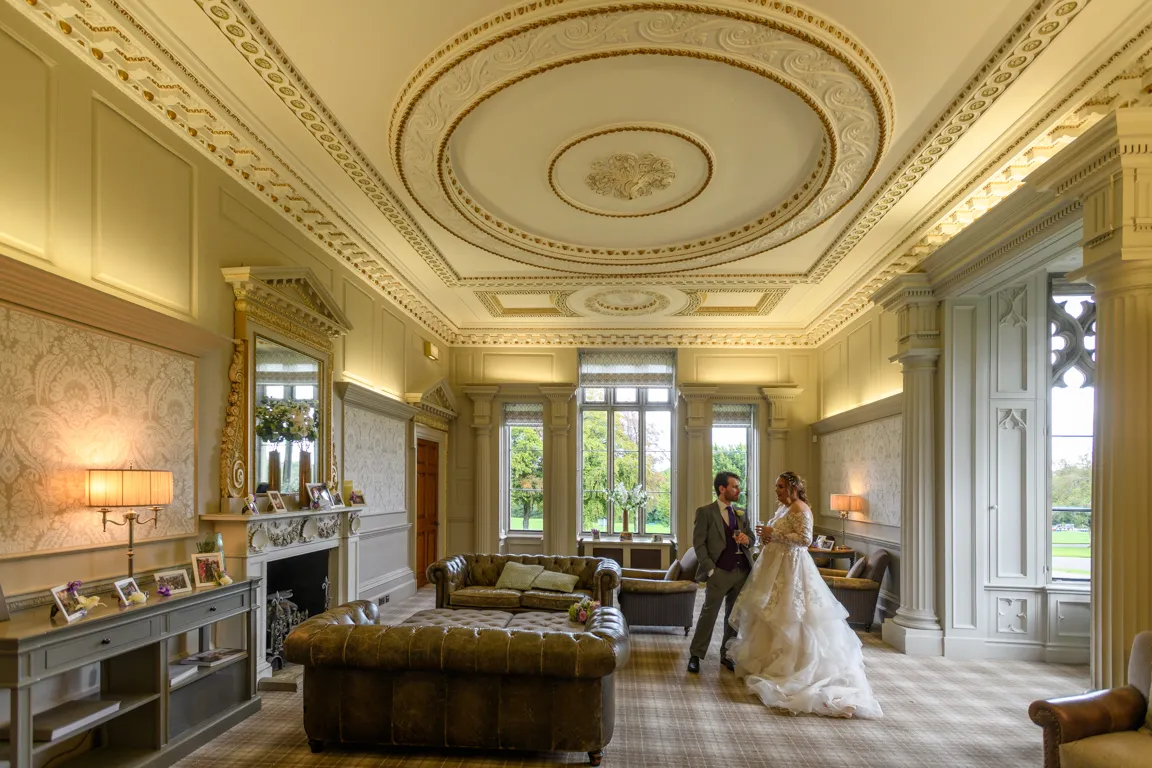 Bride in a white wedding dress and groom in a suit standing and talking inside an elegant room with ornate ceiling, leather sofas, and large windows overlooking greenery.