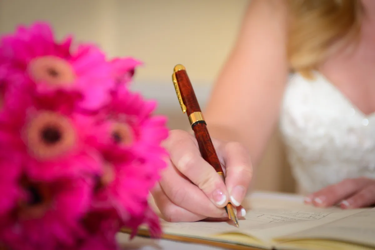 Bride signing a document with a brown and gold pen next to a pink flower bouquet.